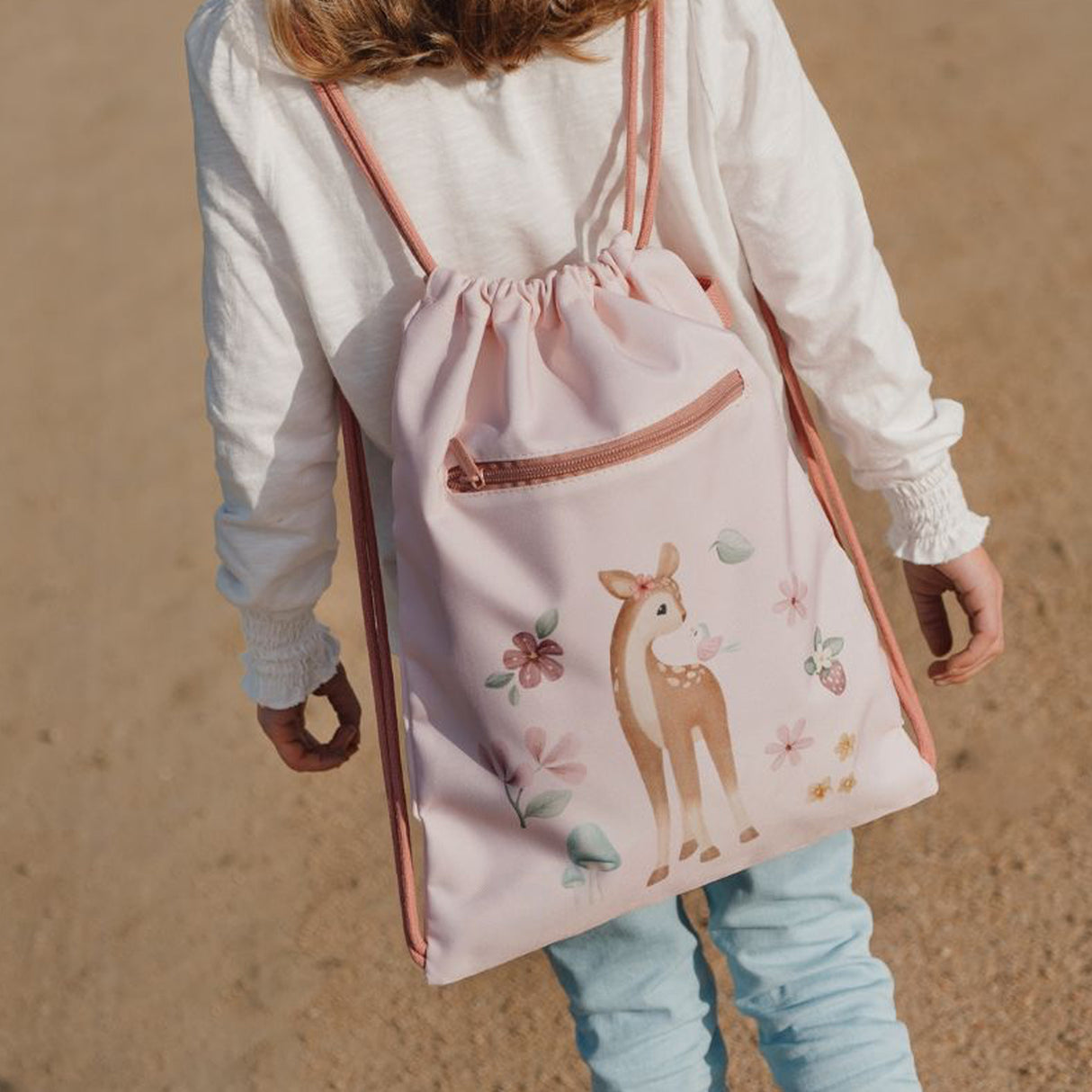 Child wearing a pink drawstring bag with a deer design on a sandy background