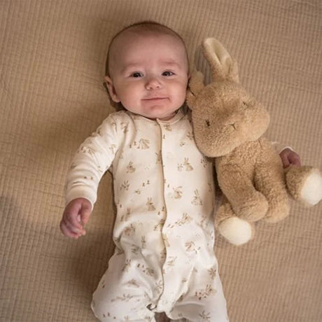 Baby in a white onesie with animal prints holding a plush bunny toy on a beige blanket.
