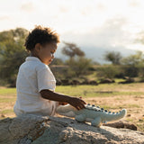 A young child sitting outdoors on a rock, touching a plush crocodile toy.