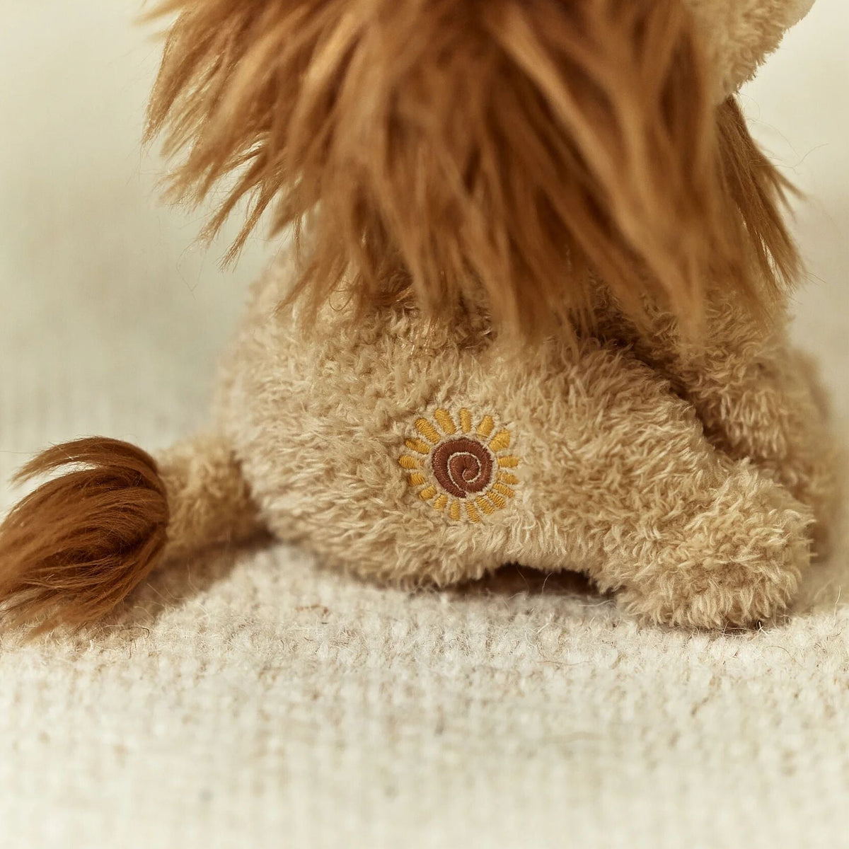 Close-up of a fluffy lion toy with a small embroidered sun detail on its side.