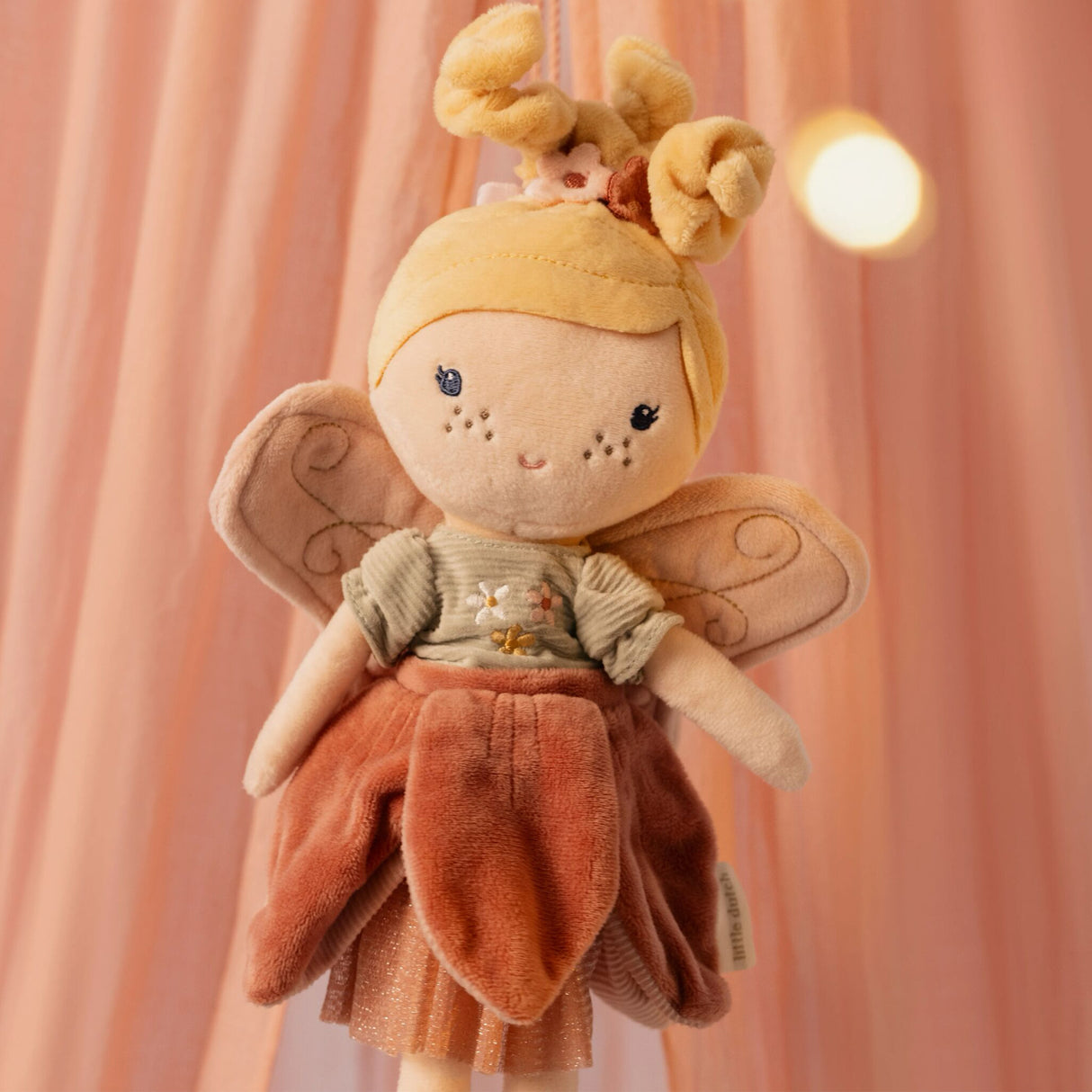 Close-up of Little Dutch Fairy Mila doll face with embroidered features and pink wings against a warm pink backdrop