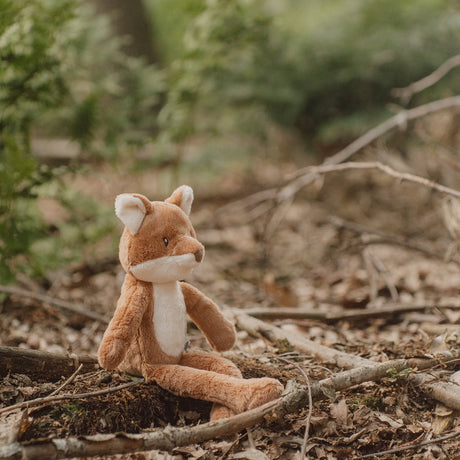 Stuffed fox toy in a natural forest setting with greenery and twigs.