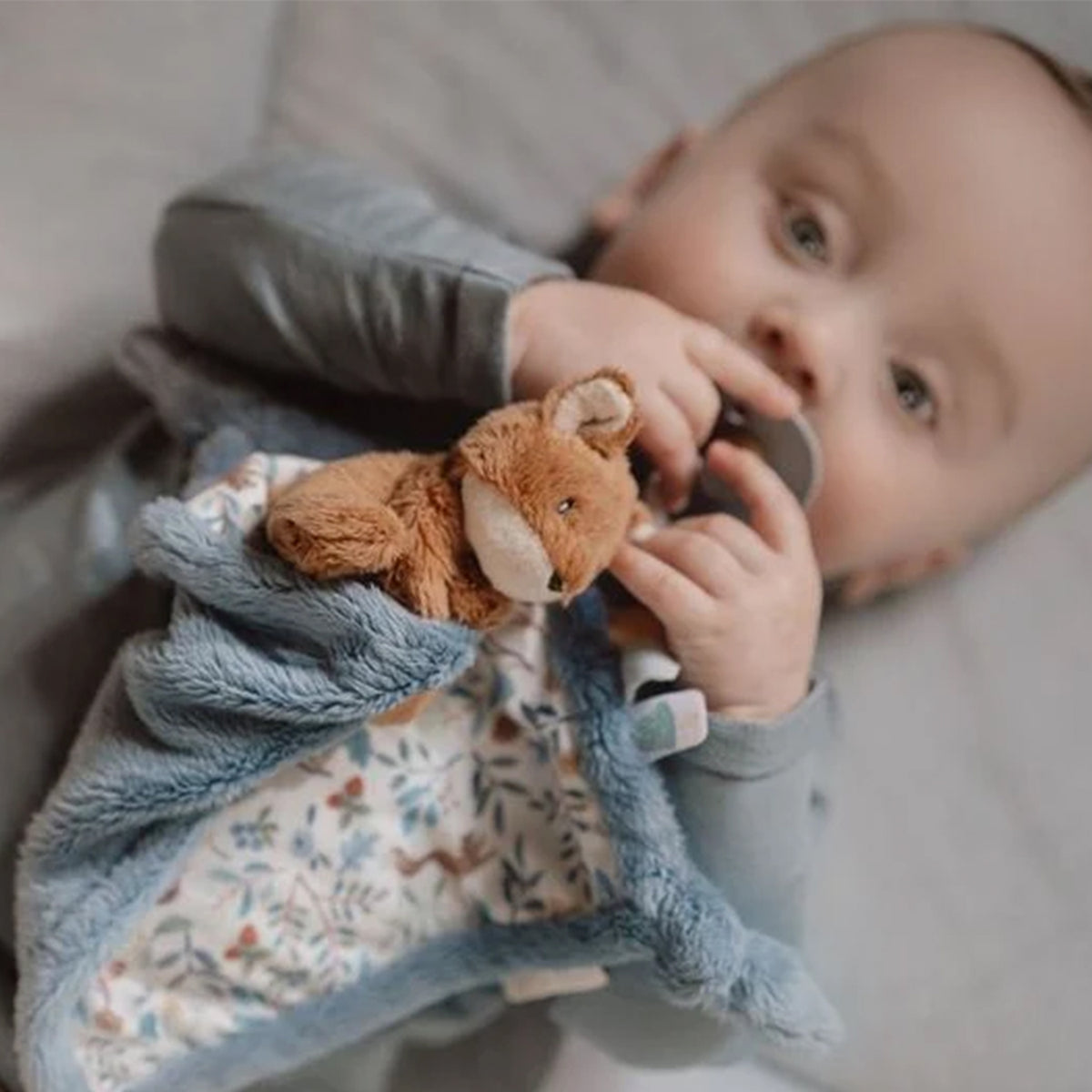 A baby lying on a soft surface, holding a small plush fox toy and a blue textured blanket with a woodland pattern.