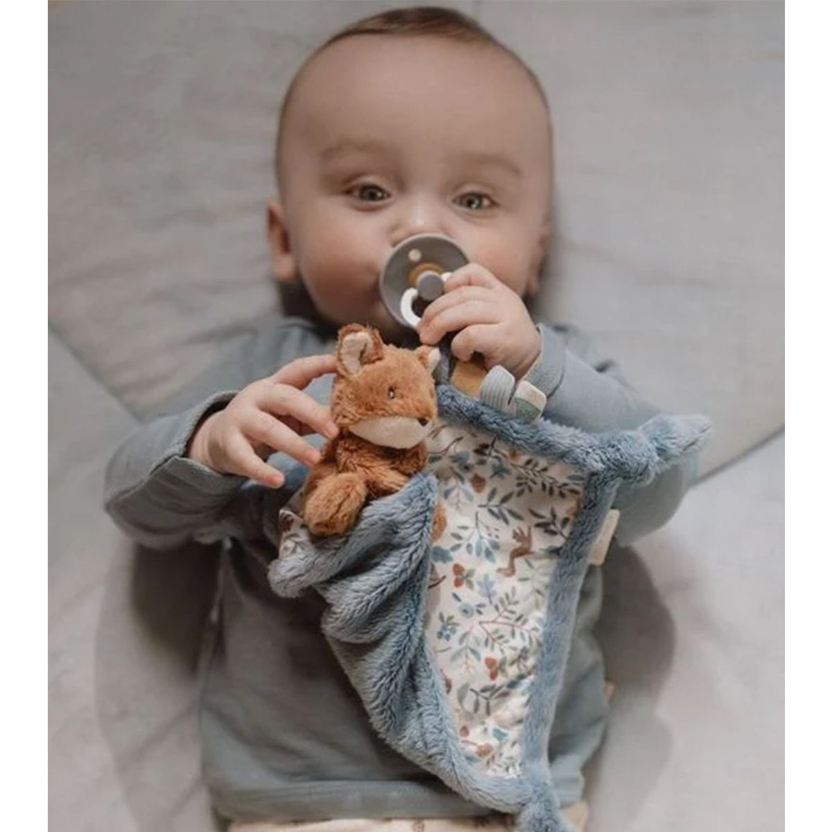 A baby lying on a bed, holding a plush fox toy attached to a blue textured sensory cloth with floral woodland blue pattern