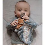 A baby lying on a bed, holding a plush fox toy attached to a blue textured sensory cloth with floral woodland blue pattern