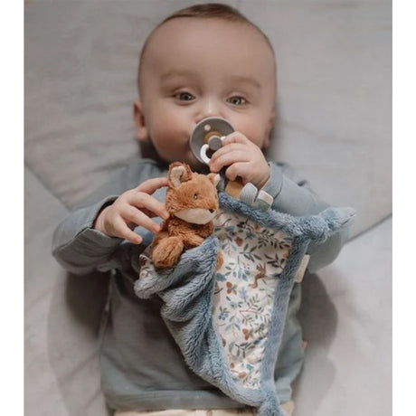 A baby lying on a bed, holding a plush fox toy attached to a blue textured sensory cloth with floral woodland blue pattern
