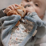 A baby holds a soft brown fox toy attached to a blue-gray plush soother blanket with a floral foxy print. 
