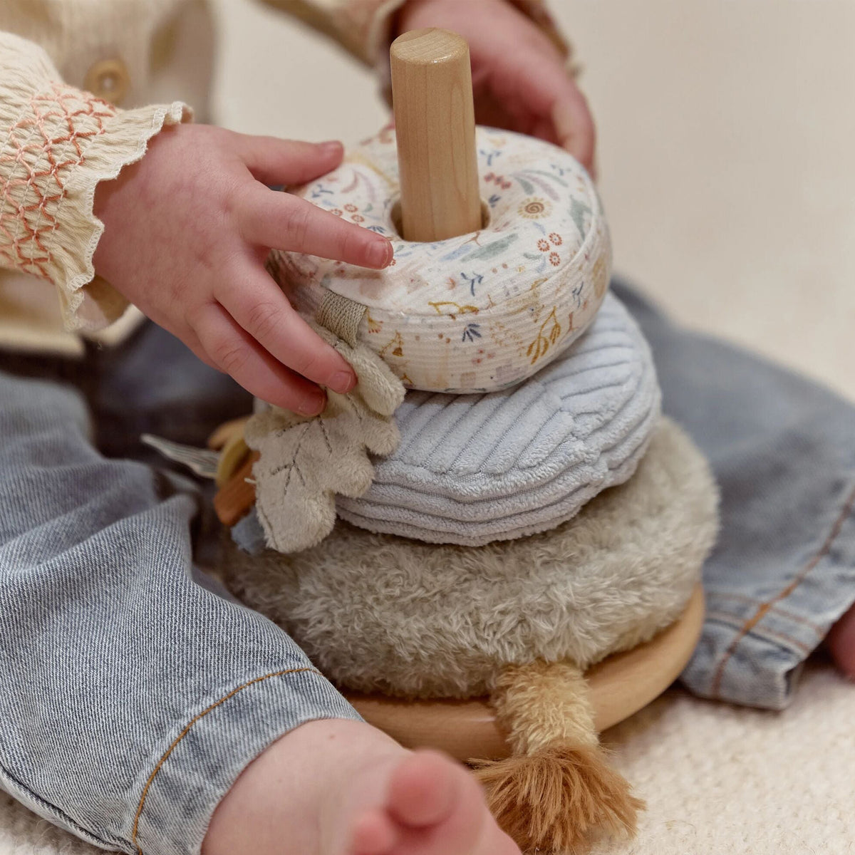 A close-up of a child's hands and legs interacting with a soft stacking toy. The toy features a light wood base and pegs, a cream fabric donut, a light blue-gray ribbed fabric ring, and a beige plush ring on a wooden base.