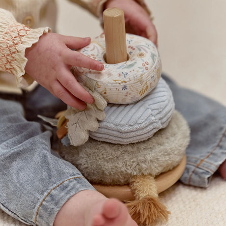 A close-up of a child's hands and legs interacting with a soft stacking toy. The toy features a light wood base and pegs, a cream fabric donut, a light blue-gray ribbed fabric ring, and a beige plush ring on a wooden base.