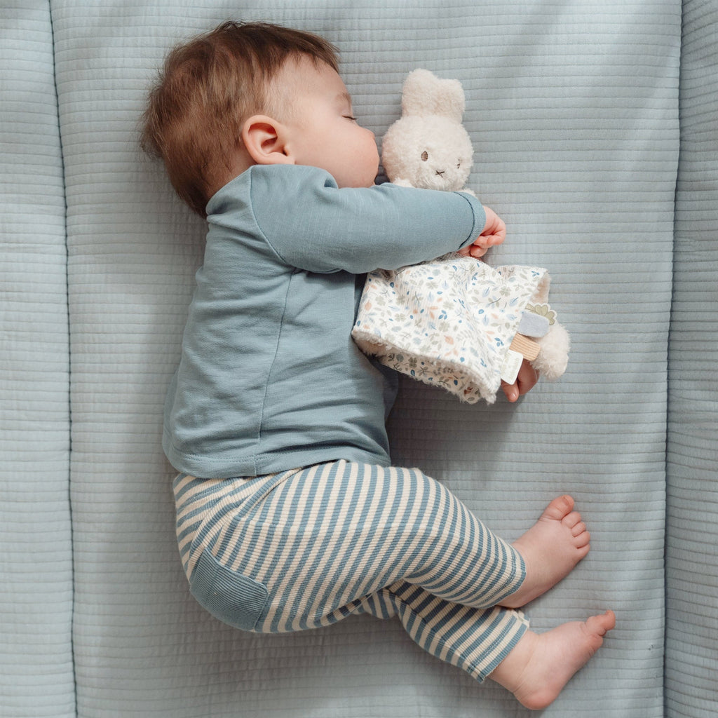 Baby in blue top and striped pants holding Miffy bunny toy with a floral cloth blanket 