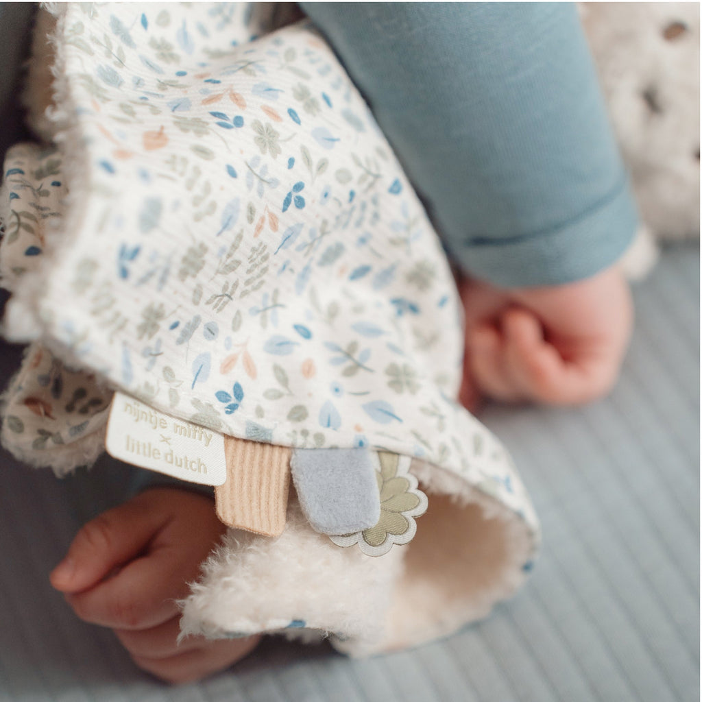 Baby in blue top and striped pants holding Miffy bunny toy with a floral cloth blanket 