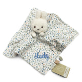 A Miffy-style security blanket with a plush white bunny attached to a cream fabric cover printed with a delicate blue, green, and peach floral pattern. A name is embroidered in blue script on the blanket