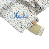 Close-up of a baby comfort blanket with a white background and a delicate blue, green, and beige floral pattern. A name is embroidered in blue script on the pattern.