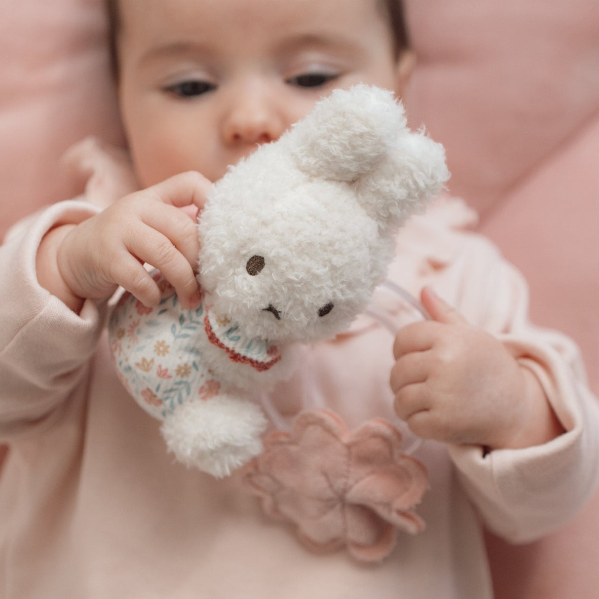 Baby holding a fluffy white Miffy ring rattle toy with a pink background