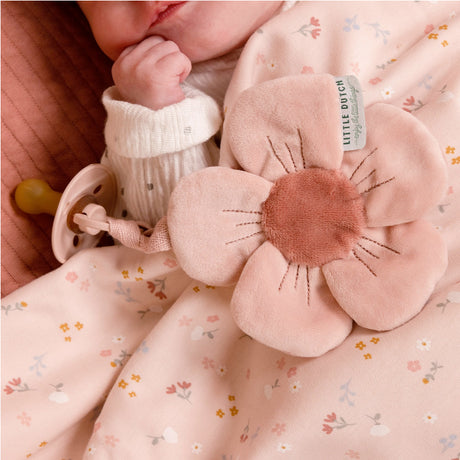 A soft pink dummy cloth shaped like a flower with an attachment, baby on bed