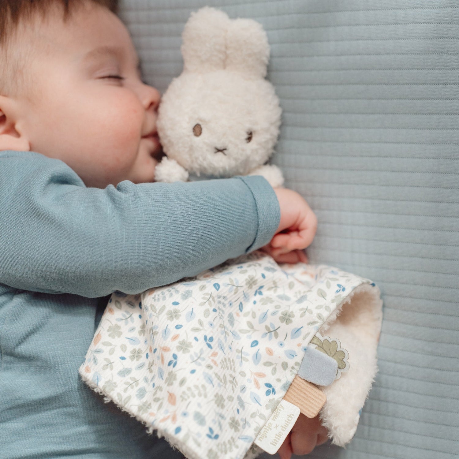 Baby in blue top and striped pants holding Miffy bunny toy with a floral cloth blanket 