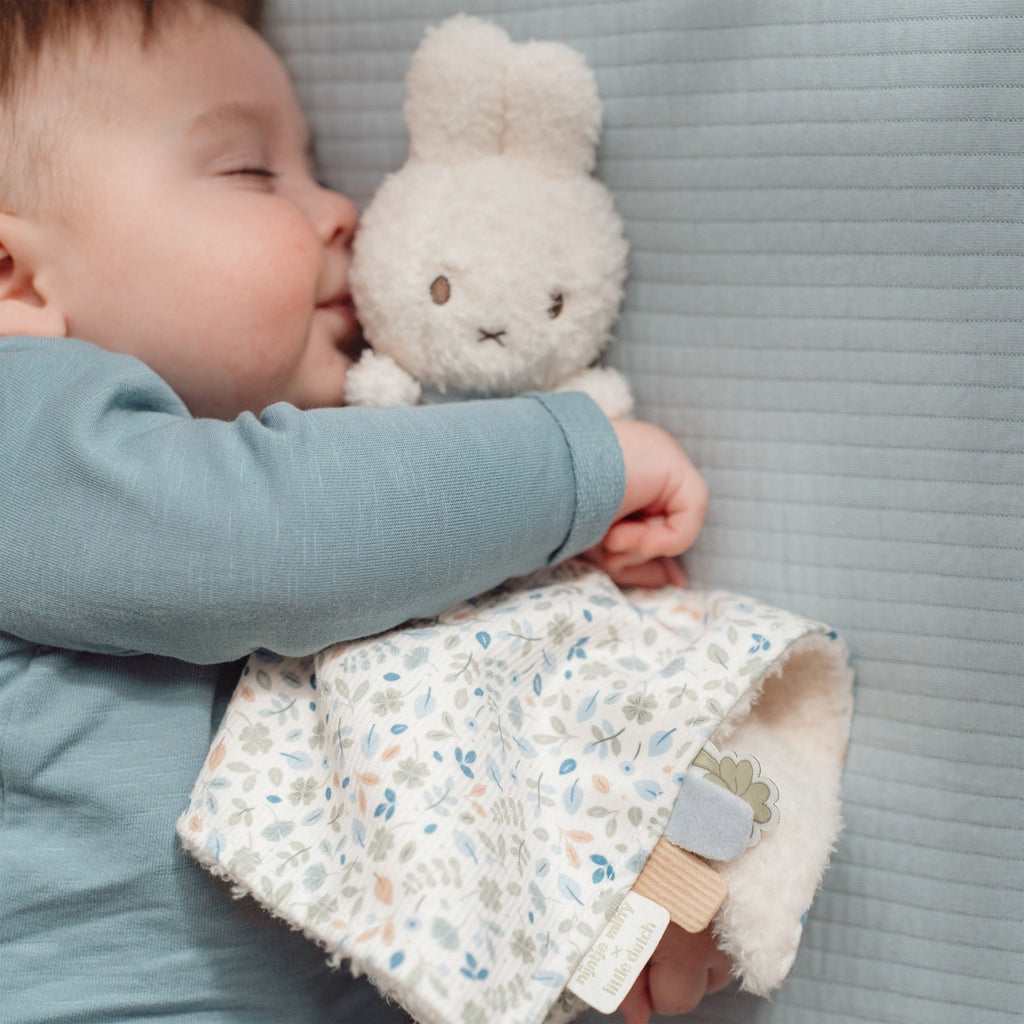 Baby in blue top and striped pants holding Miffy bunny toy with a floral cloth blanket 