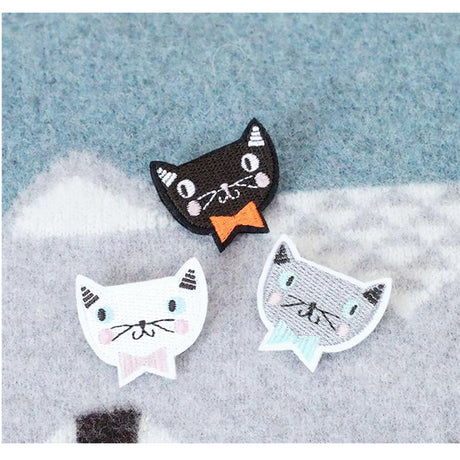 A set of three embroidered brooches in the shape of cats' faces, displayed in an open box. Each brooch has colorful details and is attached to a gold-tone safety pin.