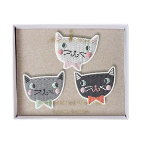 A set of three embroidered brooches in the shape of cats' faces, displayed in an open box. Each brooch has colorful details and is attached to a gold-tone safety pin.