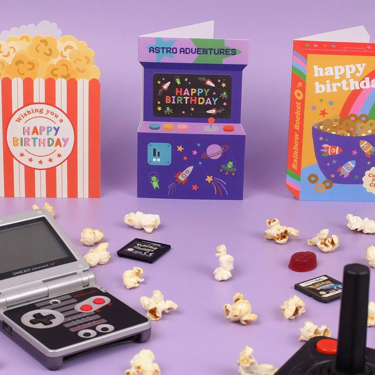 Mini arcade machine with 'Happy Birthday' card, popcorn, and retro gaming console on a purple background