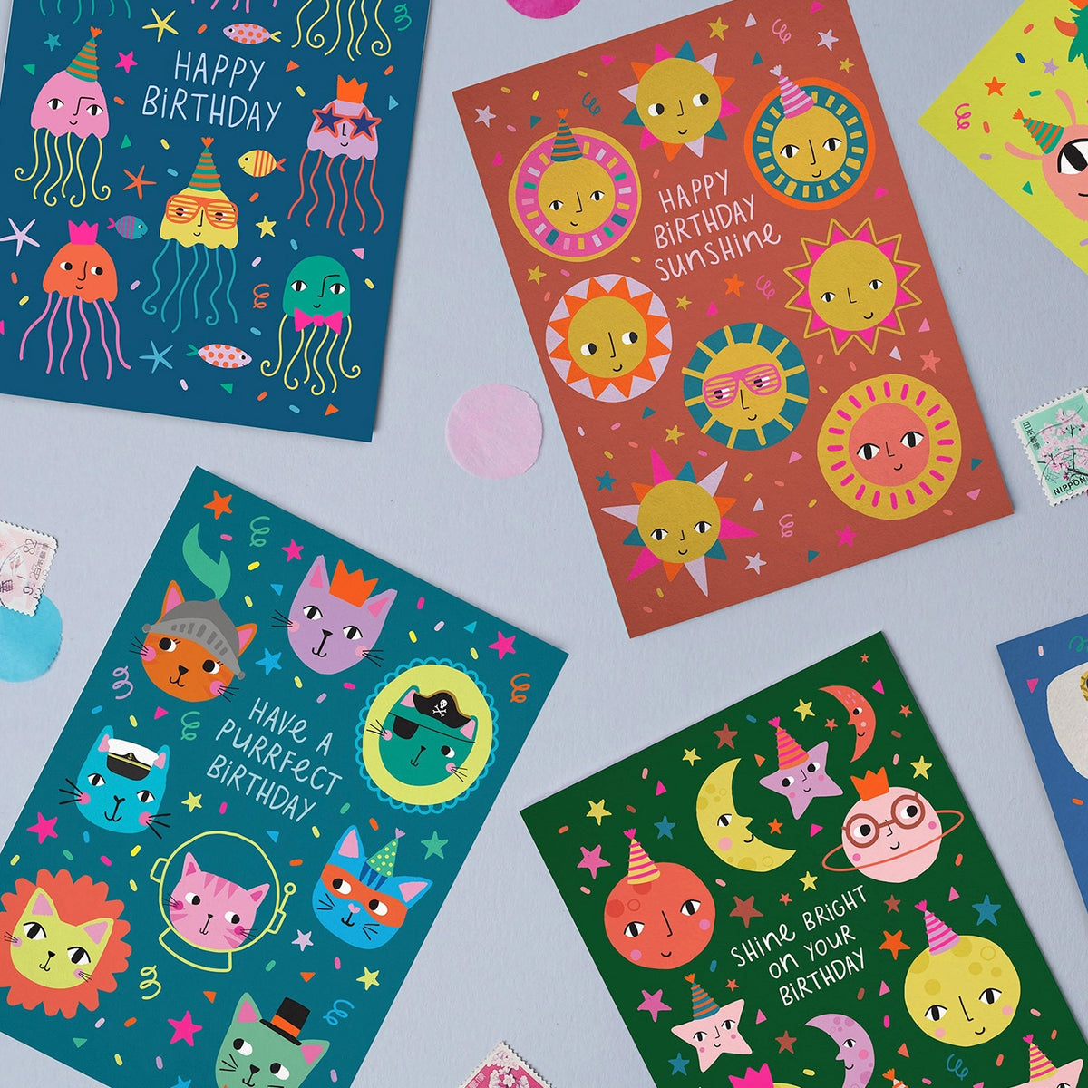 A styled flat-lay of four colourful birthday cards with cartoon illustrations, arranged on a light blue surface with small paper confetti and postage stamps.