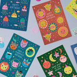 A styled flat-lay of four colourful birthday cards with cartoon illustrations, arranged on a light blue surface with small paper confetti and postage stamps.