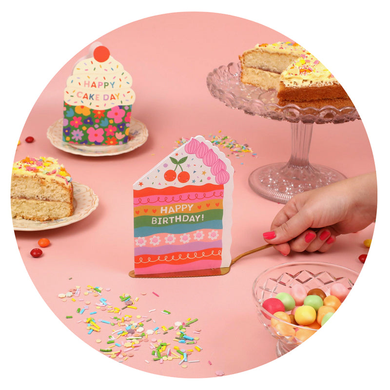 Colorful birthday cake with a small house-shaped card and a slice of cake on a pink background.