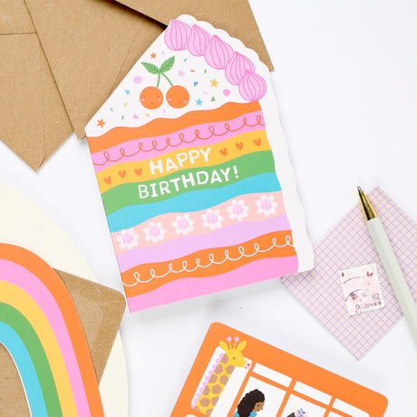 Mifkins Colorful birthday card with cake design on a white surface with stationery items.