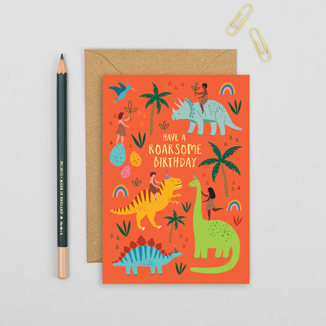 Birthday card with dinosaur illustrations on a red background, next to a pencil and paper clips.