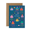 A birthday card with a brown kraft envelope behind it. The card features a dark blue background with colourful cartoon jellyfish, fish, stars, and the words 'HAPPY BIRTHDAY' in the centre.