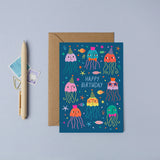 A birthday card with a brown envelope, styled on a light blue-gray surface next to a pen and two small postage stamps. The card features a dark blue underwater scene with colourful cartoon jellyfish, fish, and the words 'HAPPY BIRTHDAY'.