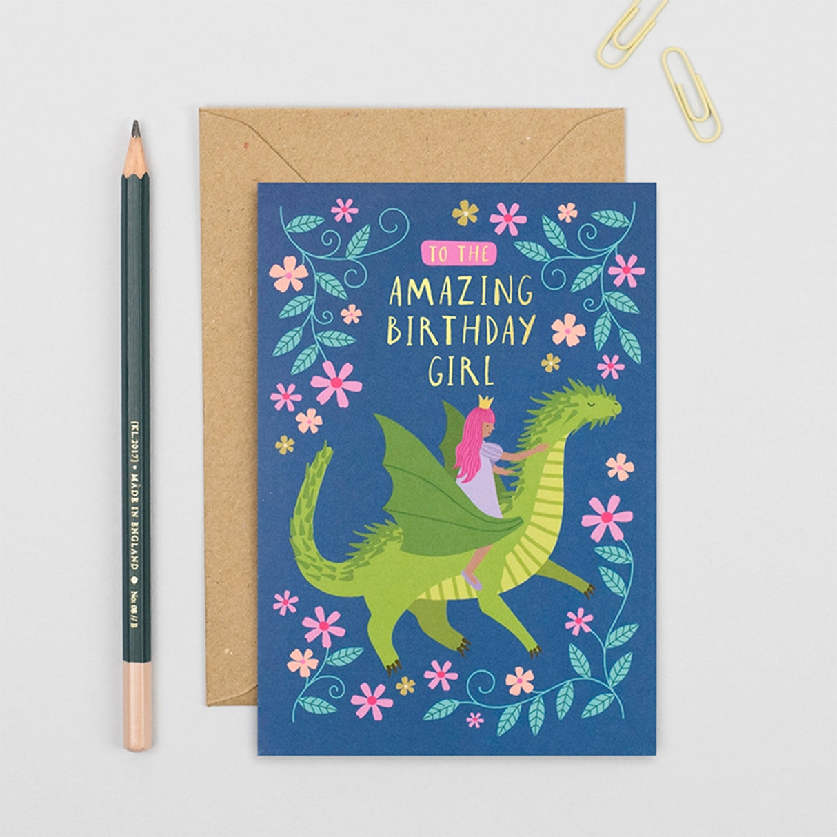 A birthday card with a blue background and an illustration of a green dragon with a pink-haired girl riding it, shown with a brown kraft envelope, a pencil, and paper clips on a light gray surface.