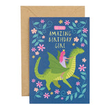 A birthday card with a kraft envelope, illustration of a green dragon and a princess riding it, surrounded by pink flowers and blue leaves on a dark blue background.