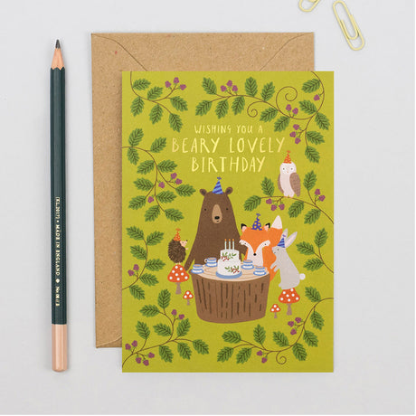 Birthday card with cartoon bears and foxes around a cake, next to a pencil on a light gray background