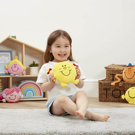 Child holding a yellow Little miss sunshine yellow soft toy in a room with toys and a shelf in the background