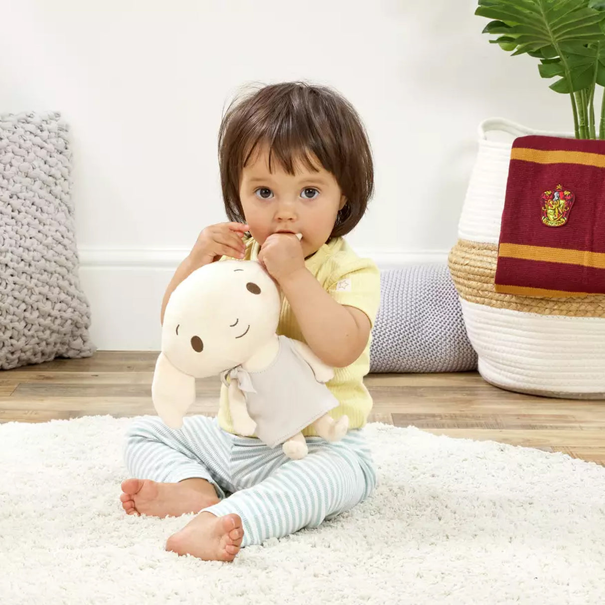 Child holding a plush soft toy Dobby character from Harry Potter. Big ears and a smiling face wearing his pillowcase. the child is sitting on a white carpet with a plant and basket in the background