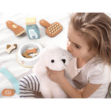 A young girl holding a white dog soft toy. Surrounding her are wooden pet care toys, including a brush, a foot, a milk carton, and a bowl with wooden bones.