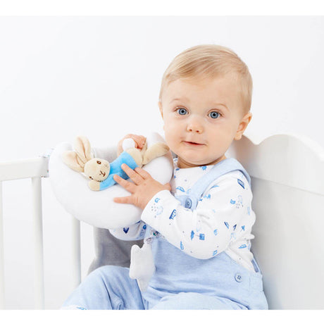 Baby sitting in a crib holding a plush Peter Rabbit musical toy with a bunny on a moon and star pull, featuring text 'Night Night' and 'Sleep Tight'. wearing a white shirt with blue patterns and light blue pants.