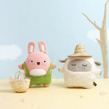 Two plush toys, a pink rabbit and a white sheep, standing on a sandy surface with a blue sky background.