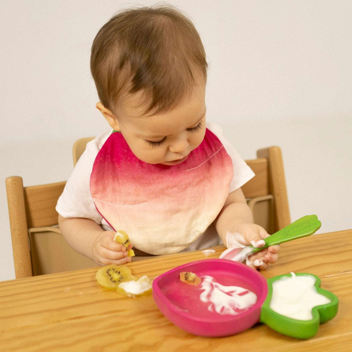 Baby sitting at a table with a colourful bib, eating food from the Oli and Carol Raddish plate and spoon set.