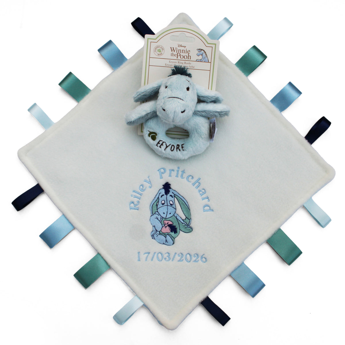 A personalized baby Taggy blanket featuring a ring rattle Eeyore toy on a cream-colored square fabric tag with ribbon tabs. The blanket is embroidered with a babies name, a cartoon Eeyore, and the date of birth.