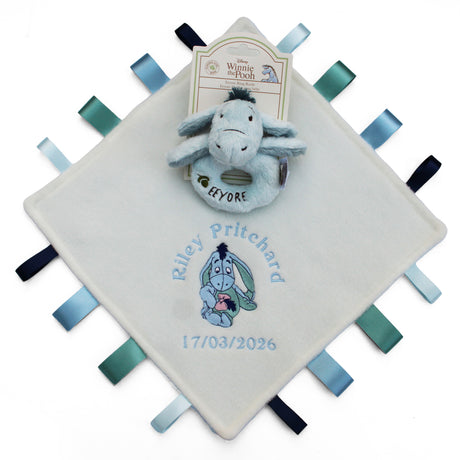 A personalized baby Taggy blanket featuring a ring rattle Eeyore toy on a cream-colored square fabric tag with ribbon tabs. The blanket is embroidered with a babies name, a cartoon Eeyore, and the date of birth.