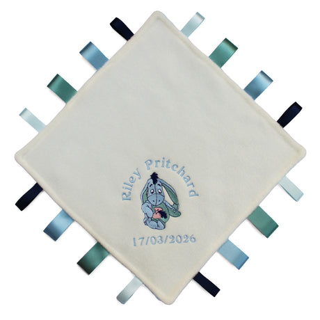 A personalised cream-coloured baby sensory lovey blanket with embroidered text and a cartoon eeyore character, surrounded by colourful ribbons on the edges.