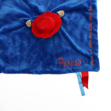 A close-up of a blue soft Paddington baby soother, with a name embroidered in red and a small red and blue ribbon.