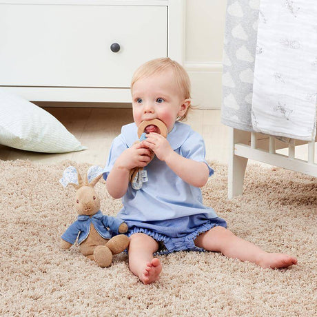Baby sitting on a carpeted floor holding a wooden toy, with a teddy bear beside them.
