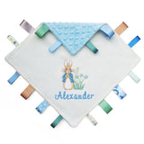 A personalized baby lovey blanket featuring an embroidered Peter Rabbit and the name 'Alexander' in blue script. The top corner is folded over to reveal a light blue textured dotty fabric lining, and multiple patterned fabric tabs in blue, green, brown, and white are stitched around the edges.