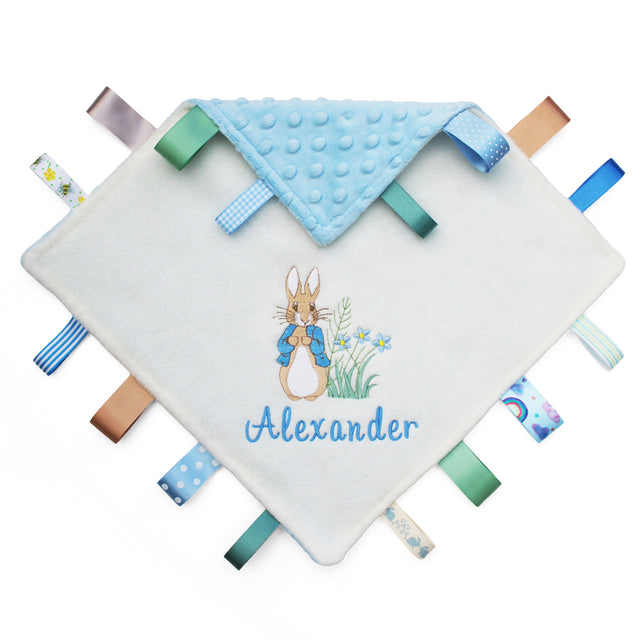 A personalized baby lovey blanket featuring an embroidered Peter Rabbit and the name 'Alexander' in blue script. The top corner is folded over to reveal a light blue textured dotty fabric lining, and multiple patterned fabric tabs in blue, green, brown, and white are stitched around the edges.