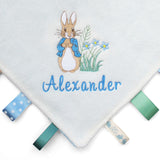 A close-up of a baby taggy blanket featuring an embroidered Peter Rabbit design, the name 'Alexander' in blue script, and colourful ribbon tabs along the edges.