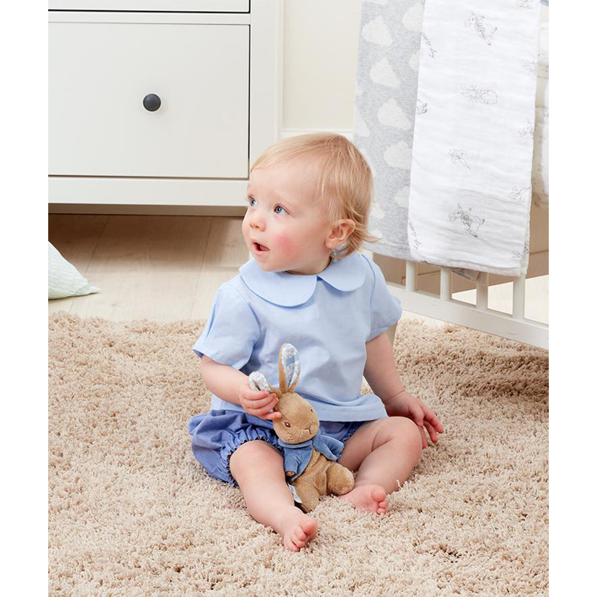 Baby in a light blue outfit sitting on a beige carpet holding a Peter Rabbit soft toy