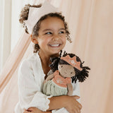 A smiling young child with curly hair holding a little dutch doll with dark curly hair, a peach crown, and a pale green dress
